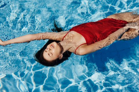 Photo for Young woman in the pool in a red swimsuit with a beautiful smile lying on the water and swimming in the sun swimming in the pool, the concept of relaxing on vacation. High quality photo - Royalty Free Image