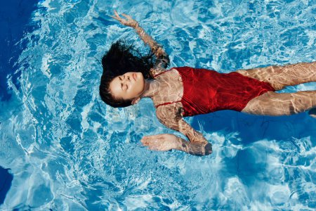 Téléchargez les photos : Jeune femme dans la piscine en maillot de bain rouge avec un beau sourire couché sur l'eau et nager au soleil dans la piscine, le concept de détente en vacances. Photo de haute qualité - en image libre de droit