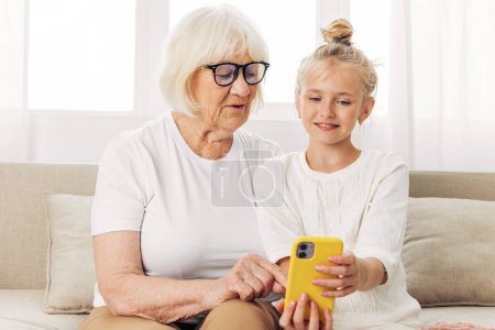Photo for People senior granddaughter indoors bonding t-shirt call education video adult space smiling selfie white two photography child copy sofa family togetherness grandmother phone happiness hugging - Royalty Free Image