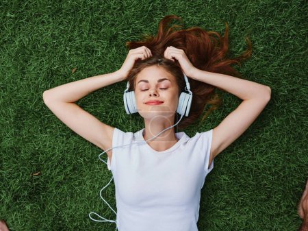Téléchargez les photos : La femme qui porte un casque écoutant de la musique repose sur une bonne humeur dans un t-shirt blanc en été sur l'herbe. Photo de haute qualité - en image libre de droit