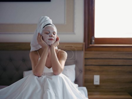Photo for Young woman lying on bed with moisturizing beauty face mask and white towel on her head after shower, lifestyle home skincare, smile. High quality photo - Royalty Free Image