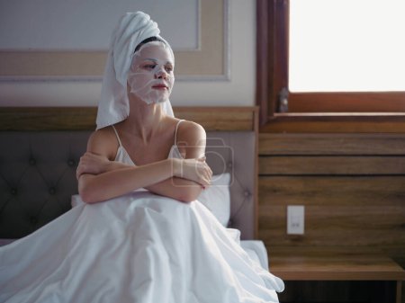 Téléchargez les photos : Jeune femme couchée sur le lit avec masque hydratant beauté et serviette blanche sur sa tête après la douche, mode de vie soins de la peau à la maison, sourire. Photo de haute qualité - en image libre de droit