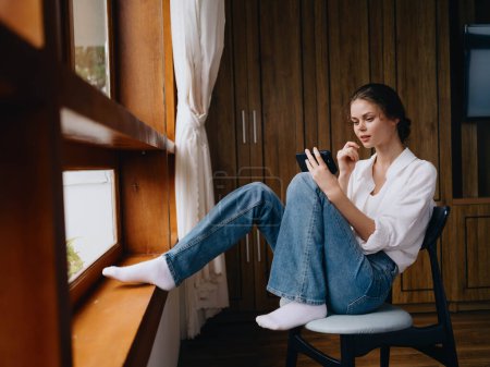Téléchargez les photos : Jeune femme assise près de la fenêtre avec téléphone dans la main sourire regardant en ligne, humeur de printemps, vêtements simples à la maison, atmosphère confortable à la maison, style de vie esthétique. Photo de haute qualité - en image libre de droit