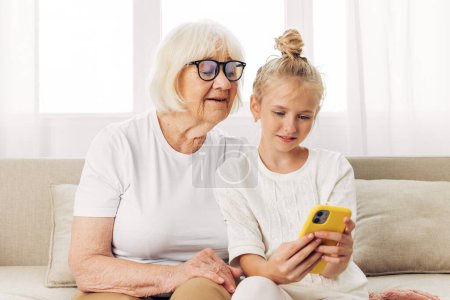 Téléchargez les photos : Éducation grand-mère selfie vidéo petite-fille canapé intérieur appel t-shirt blanc photographie espace enfant famille collage copie sourire téléphone ensemble étreinte - en image libre de droit