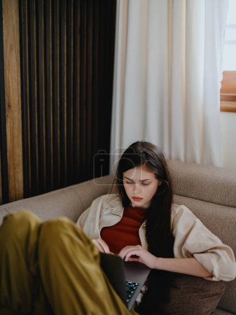 Téléchargez les photos : Une femme s'allonge sur un canapé avec un ordinateur portable et regarde pensivement à l'écran former un pigiste au travail, un vrai mode de vie sans retoucher. Photo de haute qualité - en image libre de droit