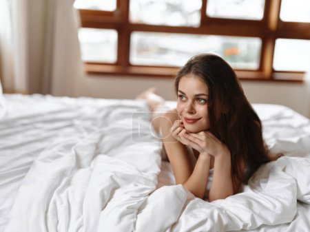 Photo for Beautiful young woman lying in bed in the morning on a day off and smiling, vacation lifestyle, homely wake up atmosphere with sunlight from the window. High quality photo - Royalty Free Image