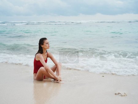Foto de Una joven sentada junto al mar en un traje de baño rojo en la arena cerca del agua y mirando al horizonte. Foto de alta calidad - Imagen libre de derechos
