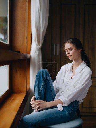 Foto de Mujer sentada en casa junto a una ventana de madera con una sonrisa en una silla en ropa cómoda casera y mirando el paisaje, el estado de ánimo de primavera, el día de las mujeres, el descanso el fin de semana. Foto de alta calidad - Imagen libre de derechos