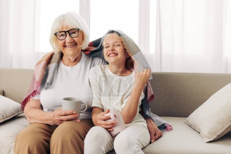 Photo for Woman granddaughter winter home love grandmother child happy couch girl plaid family hugging hug elderly old mugs sofa - Royalty Free Image