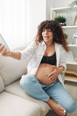 Photo for Pregnant woman blogger sits on the couch at home and takes pictures of herself on the phone, selfie and video call, consultation with the doctor online, pregnancy management. High quality photo - Royalty Free Image