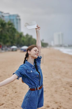 Foto de Feliz joven disfrutando de las vacaciones de verano junto al océano: una dichosa dama asiática sonriendo y de pie en una playa de arena, vistiendo un bonito vestido, con cielo azul y naturaleza tropical en el fondo. - Imagen libre de derechos