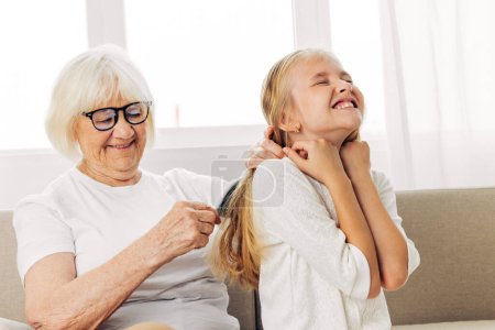 Photo for Woman girl hair family old mother grandmother sitting happy senior hug granddaughter hair home generation lifestyle couch elderly child sofa love combing comb daughter - Royalty Free Image