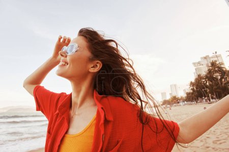 Téléchargez les photos : Serene Portrait : Une femme souriante avec des lunettes de soleil jouissant de la liberté et des couleurs vibrantes à la plage - en image libre de droit