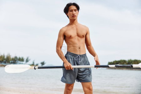 Téléchargez les photos : Homme asiatique actif profitant de l'aventure de paddleboard sur une plage d'été - en image libre de droit