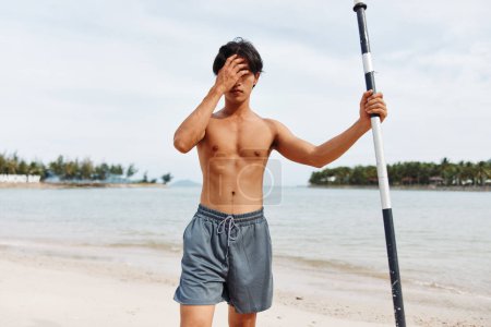 Téléchargez les photos : Homme asiatique actif profitant de vacances d'été à la plage : Paddleboard aventure dans l'océan ensoleillé - en image libre de droit