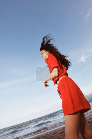 Téléchargez les photos : Femme joyeuse dansant dans la brise d'été, portant des vêtements rouges élégants, profitant de la liberté des vacances à la plage - en image libre de droit