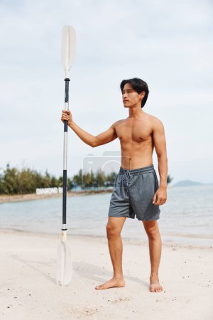 Téléchargez les photos : Beach Adventure : Homme asiatique actif faisant de l'exercice sur planche à pagaie, profitant des plaisirs de l'été et surfant sur les vagues de l'océan au coucher du soleil - en image libre de droit