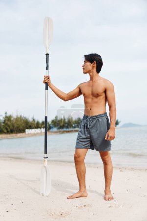 Téléchargez les photos : Homme asiatique jouissant d'un mode de vie actif sur une plage d'été avec une planche à pagaie, éprouver de l'excitation et de la sérénité au milieu du bleu vibrant de l'océan et du ciel - en image libre de droit