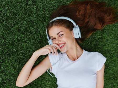 Foto de Hermosa mujer con auriculares escuchando música tumbada en la hierba en el césped en el parque en primavera y sonriendo con dientes. Foto de alta calidad - Imagen libre de derechos