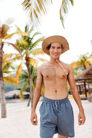 Téléchargez les photos : Homme asiatique musclé souriant sur une plage tropicale, embrassant les vibrations estivales avec des palmiers et de l'eau claire de l'océan bleu - en image libre de droit