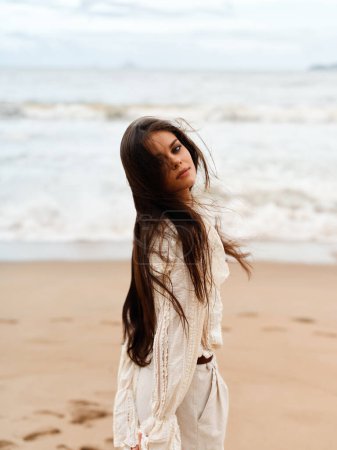 Téléchargez les photos : Beauté de l'été : Portrait attrayant des jeunes femmes, Modèle de mode en robe blanche, Debout sur la plage, Sous le ciel bleu ensoleillé, avec du vent dans ses cheveux - en image libre de droit