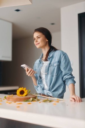 Foto de Conversaciones conectadas: cautivando a la mujer en la cocina, sosteniendo un teléfono inteligente, participando en una videollamada matutina mientras disfruta de una comida saludable en un hogar moderno. - Imagen libre de derechos