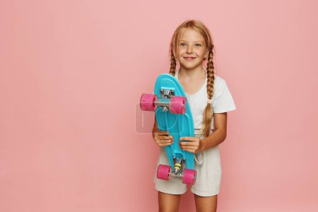 Téléchargez les photos : Joyeuse petite patineuse chevauchant une planche de Penny rose dans un portrait d'été élégant - en image libre de droit