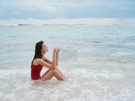 Téléchargez les photos : Femme avec un beau touriste bronzé dans un maillot de bain rouge assis sur le sable sur la plage dans l'océan dans les vagues coûteuses. Photo de haute qualité - en image libre de droit