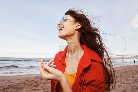 Téléchargez les photos : Femme souriante embrassant la liberté : Portrait d'une femme colorée et branchée aux lunettes de soleil bénéficiant de la vue grand angle sur l'océan au coucher du soleil sur une belle plage - en image libre de droit