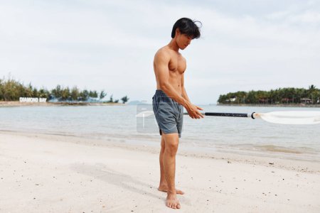Téléchargez les photos : Homme asiatique actif Profitant des plaisirs de l'été, pagayant sur une planche à pagaie debout au coucher du soleil sur une plage, entouré par la beauté de la nature et de l'océan. - en image libre de droit