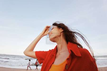 Foto de Mujer sonriente disfrutando de la libertad de una colorida puesta de sol junto al mar, con gafas de sol y ropa de playa roja de moda - Imagen libre de derechos