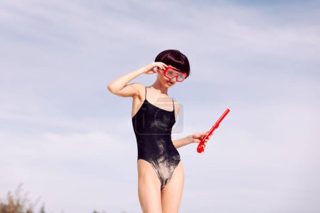Foto de Mujer sonriente haciendo snorkel en un traje de baño de moda roja, con una máscara de tema tropical y mirando a la cámara con alegría - Imagen libre de derechos