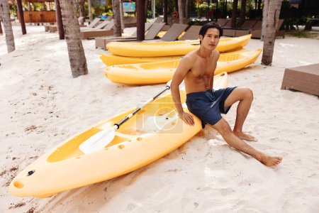 Téléchargez les photos : Serene Seascape : Un homme asiatique heureux faisant du kayak sur une plage tropicale, profitant de vacances d'été saines et actives - en image libre de droit