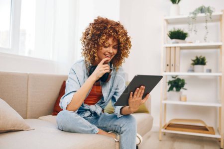 Foto de Happy Woman Holding Tablet PC y auriculares en el sofá, lectura y escucha de música en un hogar acogedor - Imagen libre de derechos