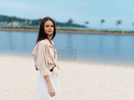 Téléchargez les photos : Jeune dame élégante, seule au bord de l'océan, profitant de vacances d'été relaxantes sur une plage, regardant élégant et heureux sous le ciel bleu et la lumière du soleil blanc - en image libre de droit