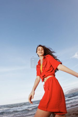 Foto de Bailando con alegría: Mujer sonriente abrazando la libertad en una moda de playa roja, disfrutando de la diversión de verano junto al mar azul - Imagen libre de derechos