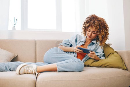 Foto de Mujer feliz sentada en un acogedor sofá en casa, sosteniendo su smartphone y sonriendo mientras disfruta de juegos en línea y leyendo mensajes en un ambiente relajado y alegre - Imagen libre de derechos