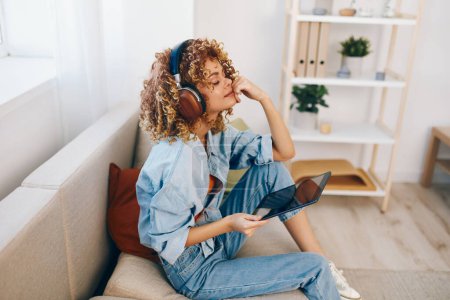 Foto de Acogedora vida: Mujer feliz leyendo, escuchando música y charlando en línea en el sofá con la tableta y los auriculares - Imagen libre de derechos