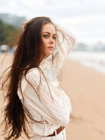 Téléchargez les photos : Serene Summer Beauty : Une jeune femme séduisante posant en plein air sur une plage de sable blanc au bord de l'océan calme, ses cheveux bruns coulant dans le vent, portant un bikini élégant sous le ciel bleu ensoleillé - en image libre de droit