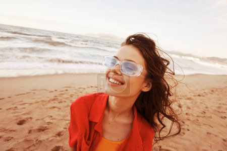 Téléchargez les photos : Femme souriante aux lunettes de soleil colorées, jouissant de la liberté et de la beauté de l'océan au coucher du soleil. - en image libre de droit