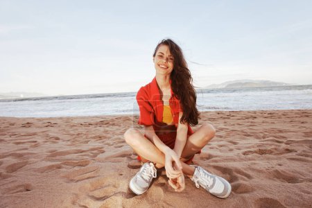 Téléchargez les photos : Femme joyeuse assise sur la plage de sable, profitant de vacances et souriant large en tenue de mode à la mode - en image libre de droit