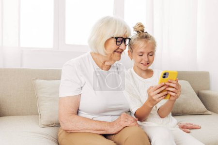 Téléchargez les photos : Grand-mère selfie ensemble espace enfant téléphone vidéo appel t-shirt canapé photographie blanc collage petite-fille étreinte à l'intérieur éducation familiale copie souriante - en image libre de droit