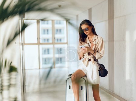Foto de Esperando: Joven viajera sentada en la terminal del aeropuerto con su equipaje y usando un smartphone, esperando su vuelo. - Imagen libre de derechos