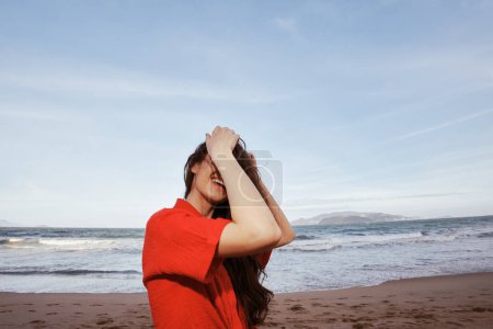 Téléchargez les photos : Liberté et joie : Une femme souriante embrassant la mer en vêtements rouges sur une plage d'été - en image libre de droit