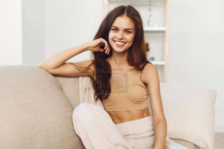 Téléchargez les photos : Femme heureuse assise sur un canapé confortable, regardant la caméra avec un sourire confiant et joyeux dans un intérieur moderne et blanc - en image libre de droit