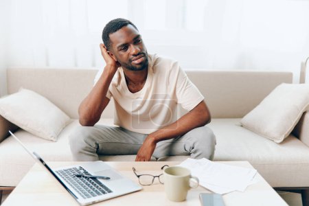 Photo for Tired African American Freelancer Working on Laptop in a Modern Home Office, Expressing Sadness and Unhappiness The man is sitting on a sofa in his living room, surrounded by cyberspace and technology - Royalty Free Image