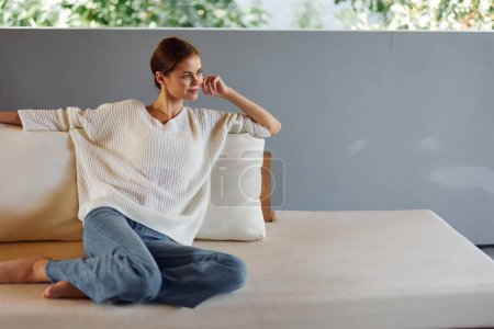 Photo for Happy Woman Sitting on Cozy Couch at Home, Relaxing and Smiling In this beautiful portrait, a young woman is seen sitting comfortably on a cozy couch in her apartment She wears a warm sweater and has - Royalty Free Image