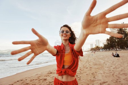 Foto de Puesta de sol colorida: Mujer sonriente con gafas de sol de moda y boca abierta que expresa alegría y libertad en la playa, manos en la arena: unas vacaciones de verano felices y despreocupadas - Imagen libre de derechos