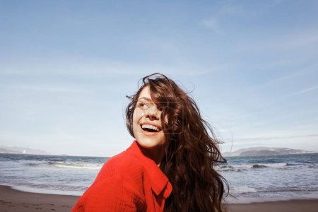 Téléchargez les photos : Joyeuse femme libérée par la mer : Émotion souriante dans des vêtements rouges pendant les vacances à la plage - en image libre de droit