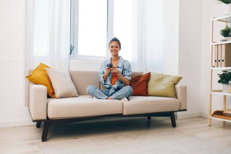 Téléchargez les photos : Femme souriante tenant un téléphone portable sur un canapé confortable dans le salon - en image libre de droit